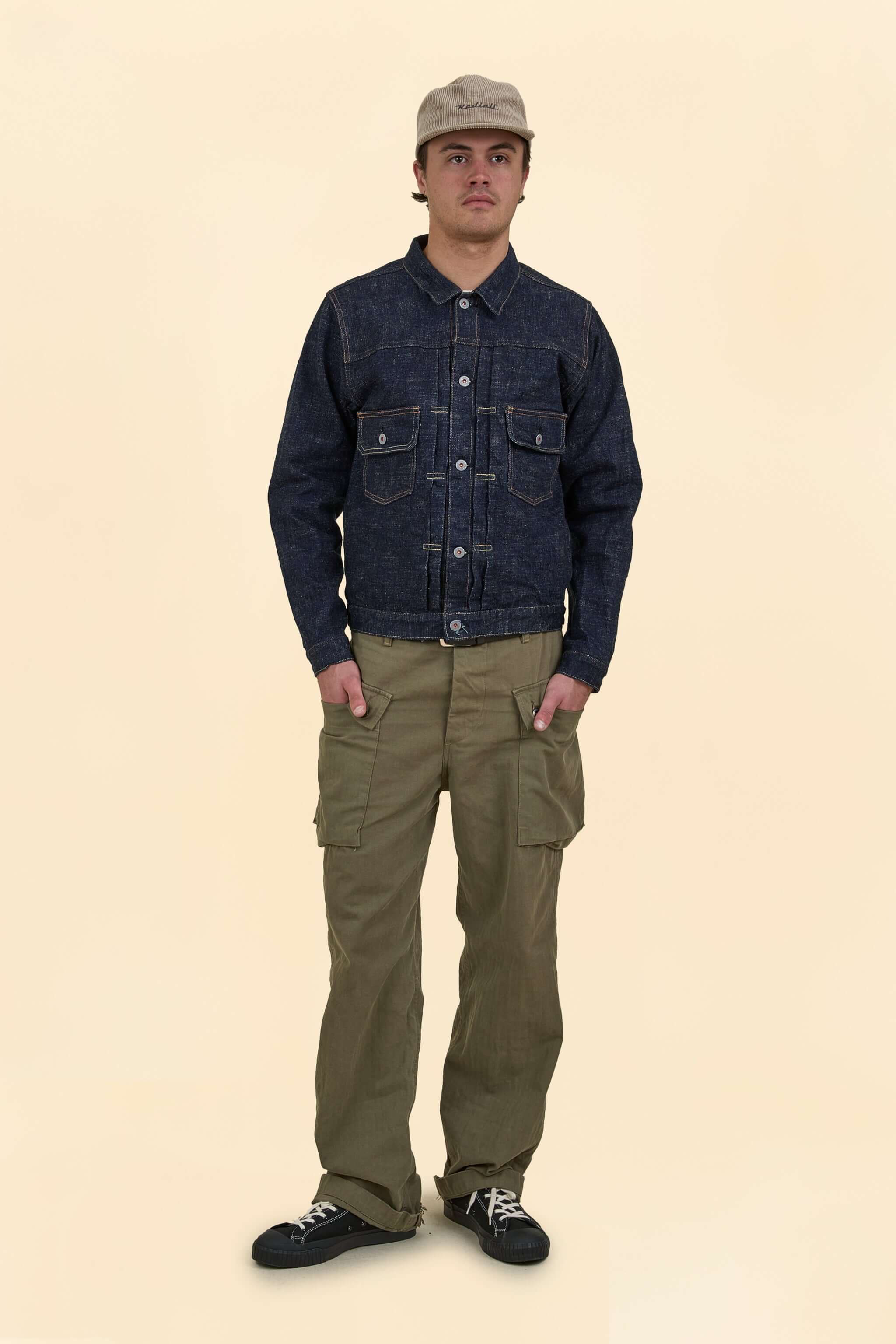 Man wearing a dark denim jacket and olive green pants on a beige background